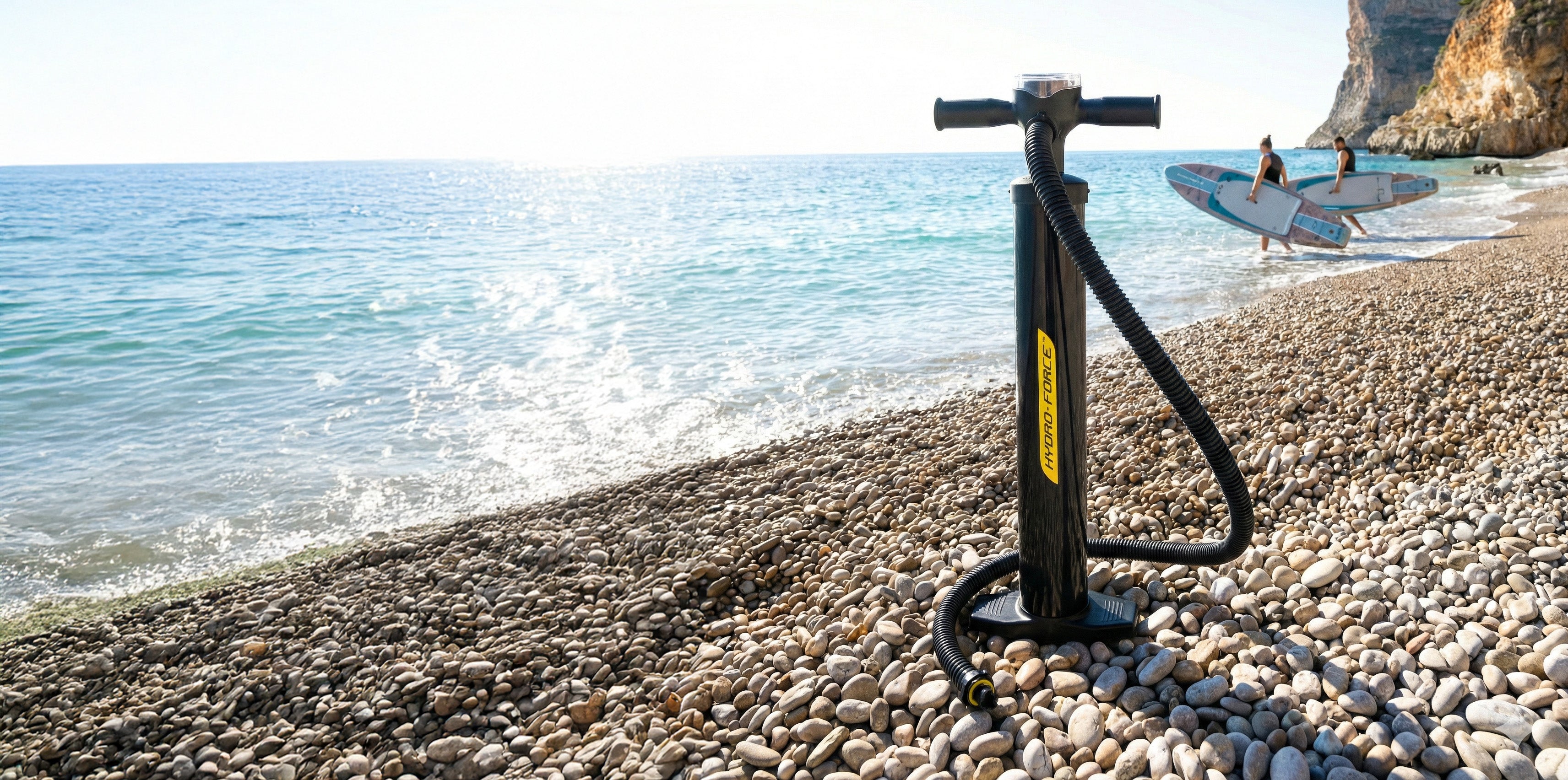 A Hydro Force air pump sitting on rocks on the shoreline with sparkling water and two paddleboarders in the background.